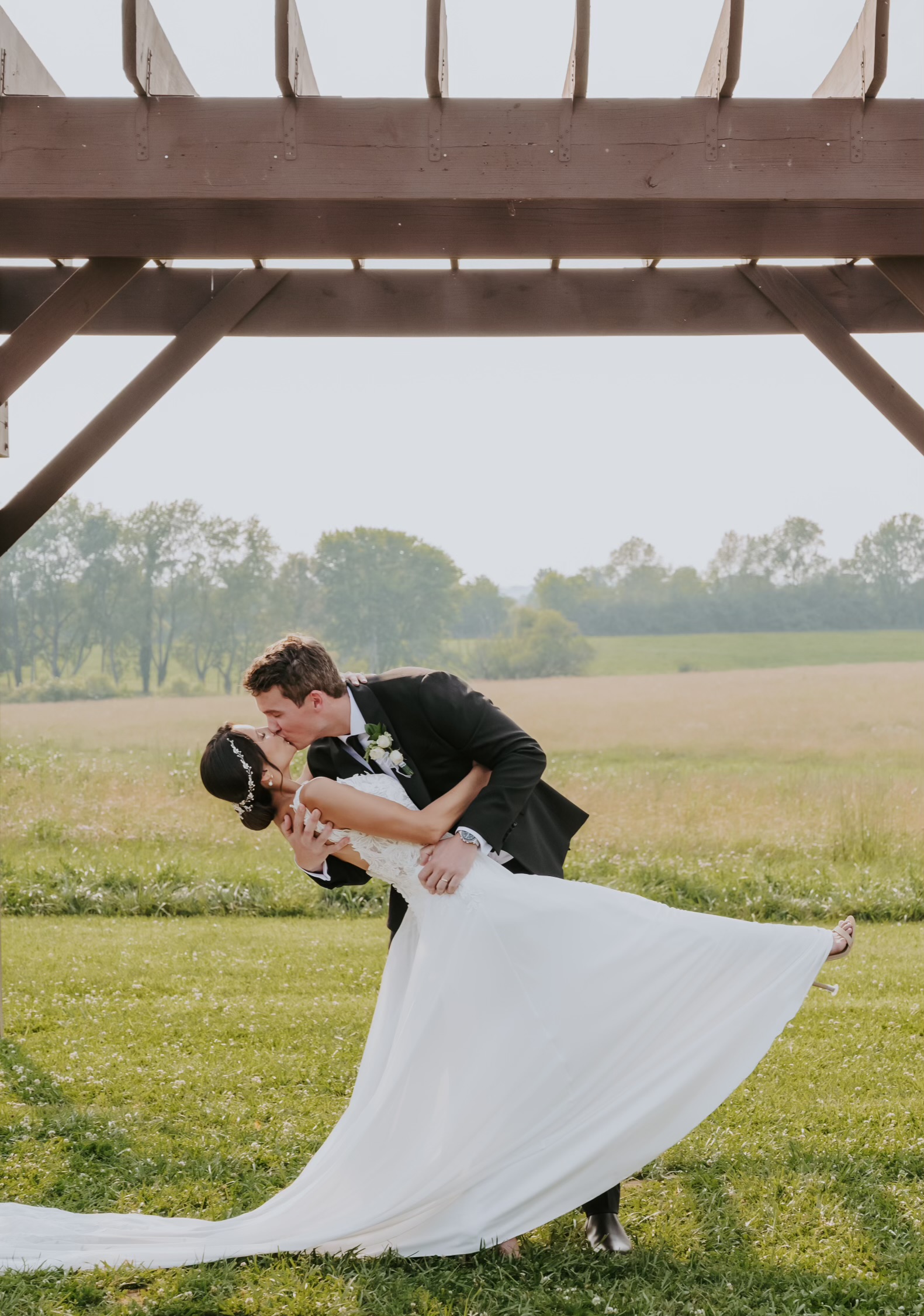 Groom dipping bride for a kiss under rustic pergola in open field